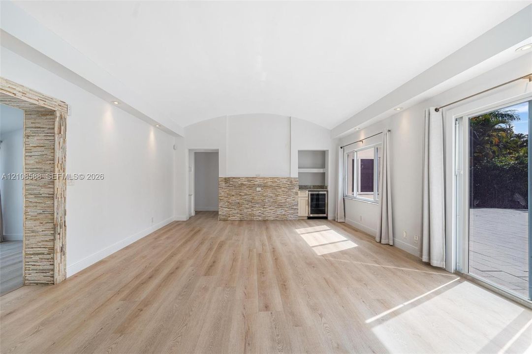 Vaulted ceiling featuring recessed lighting, stacked stone accent wall, integrated beverage refrigerator, and wood-finish flooring with sliding glass door access to outdoor hardscaping