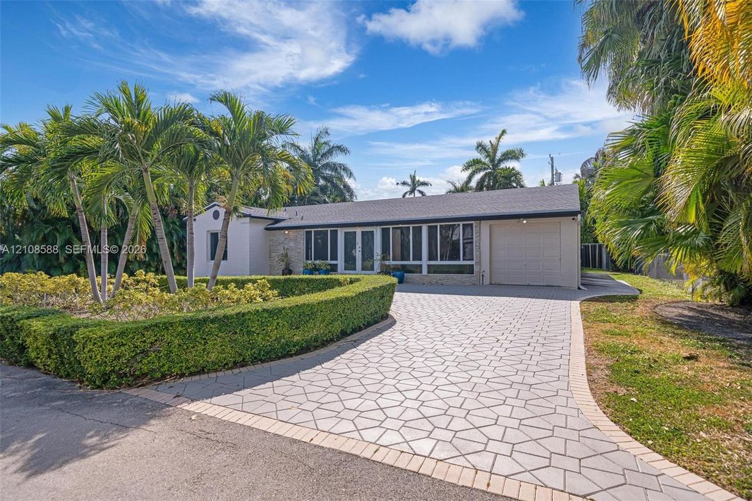 Single-story residence featuring a paved patterned driveway, attached garage, stone-accented facade, and manicured hedge landscaping with mature palm trees