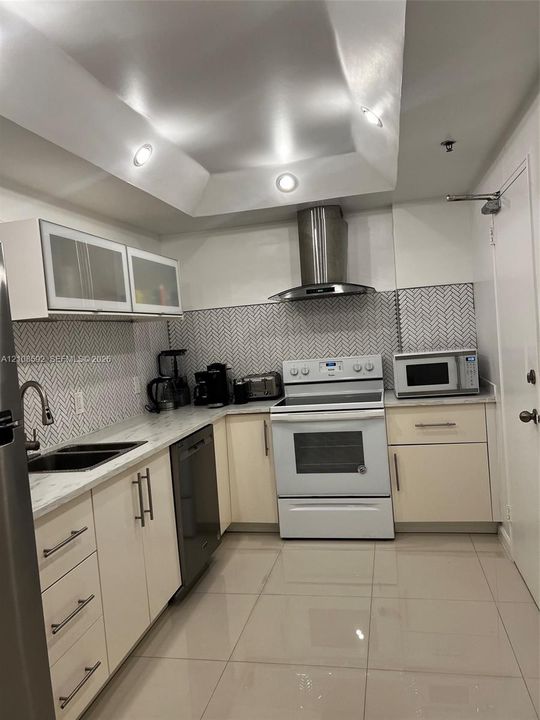 Kitchen featuring high-gloss cabinetry, stone-finish countertops, herringbone-patterned tile backsplash, stainless steel chimney-style range hood, and recessed tray ceiling lighting