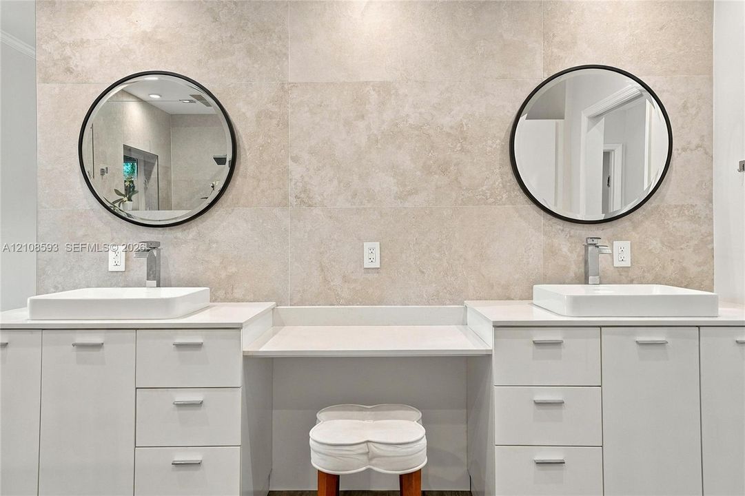 Dual white cabinetry with white stone countertops, integrated center vanity station, dual vessel sinks, wall-mounted round mirrors with matte black frames, and neutral tile wall surround