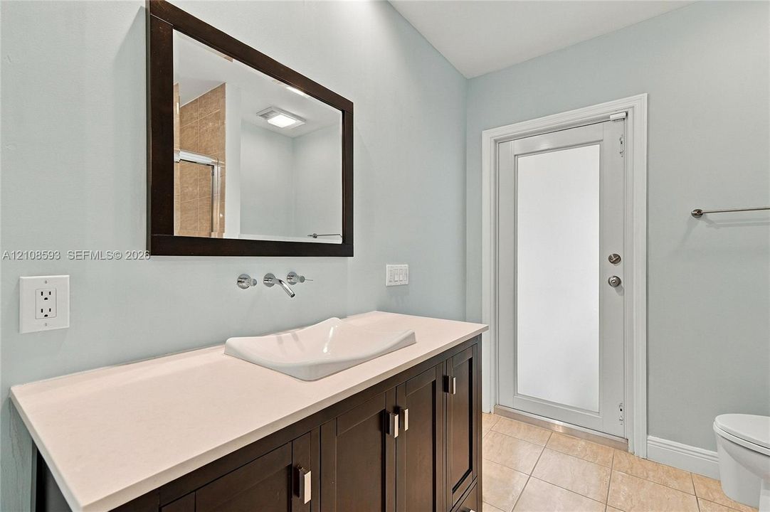 Vanity featuring a stone-surface countertop, rectangular vessel sink, wall-mounted faucet, framed wall mirror, and dark-finish cabinetry with metallic hardware, paired with a door featuring a frosted glass insert