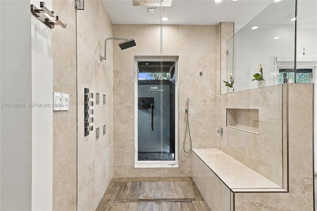 Walk-in shower featuring large-format neutral stone wall tile, multi-head shower system with a rain shower head, built-in stone bench with integrated recessed shelving, and wood-finish flooring