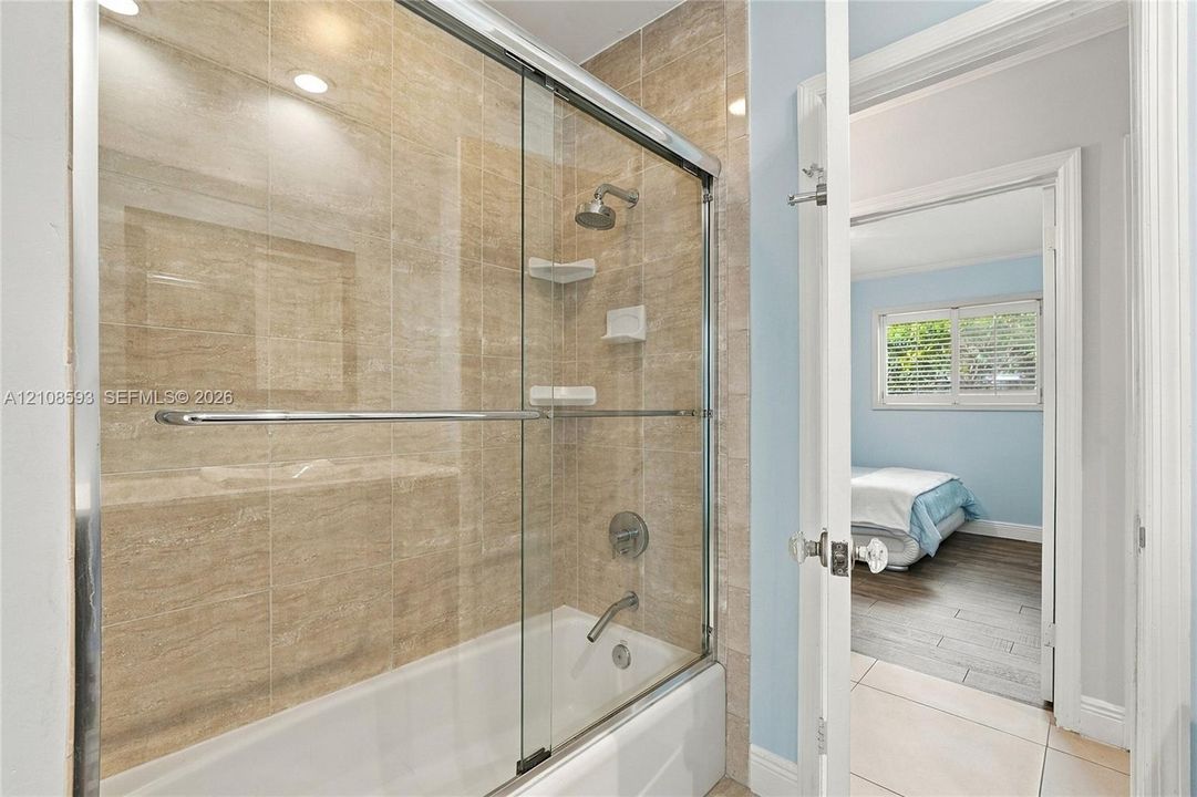 Bathtub and shower combination with floor-to-ceiling neutral tone tile surround, sliding glass enclosure, integrated corner shelving, and chrome faucet fixtures