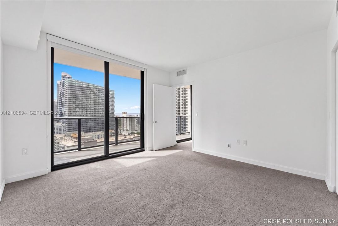 Floor-to-ceiling glass sliding doors, private balcony with metal railing, carpeted flooring, and urban cityscape scenery