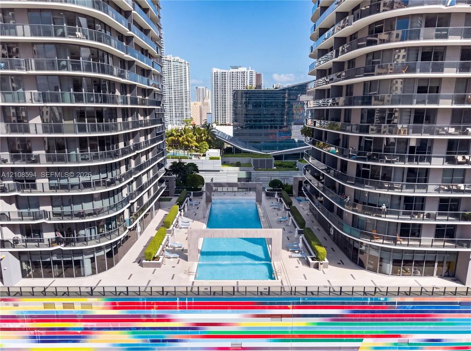 Outdoor pool featuring concrete deck surfaces, decorative stone archways, manicured hedges, and multi-story high-rise buildings with glass-paneled balconies