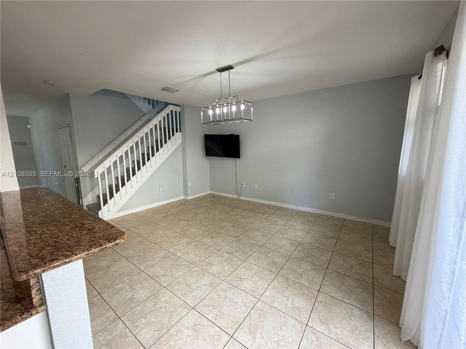 Open-concept interior featuring large-format tile flooring, stone-surfaced peninsula countertop, staircase with white balusters, and a rectangular multi-light chandelier