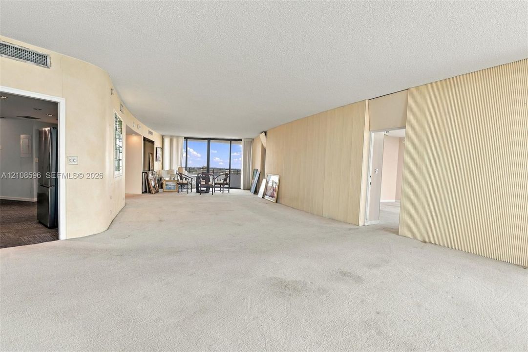 Open-concept floor plan featuring expansive light-toned wall paneling, curved wall architecture, floor-to-ceiling glass sliding doors, and neutral wall-to-wall carpeting
