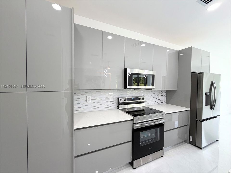 Modern kitchen featuring high-gloss gray cabinetry, white solid-surface countertops, a mosaic tile backsplash, and stainless steel appliances