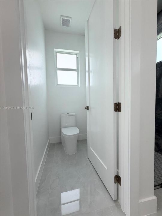 Elongated white toilet, large-format neutral tile flooring, and a single-panel interior door with dark metal hardware
