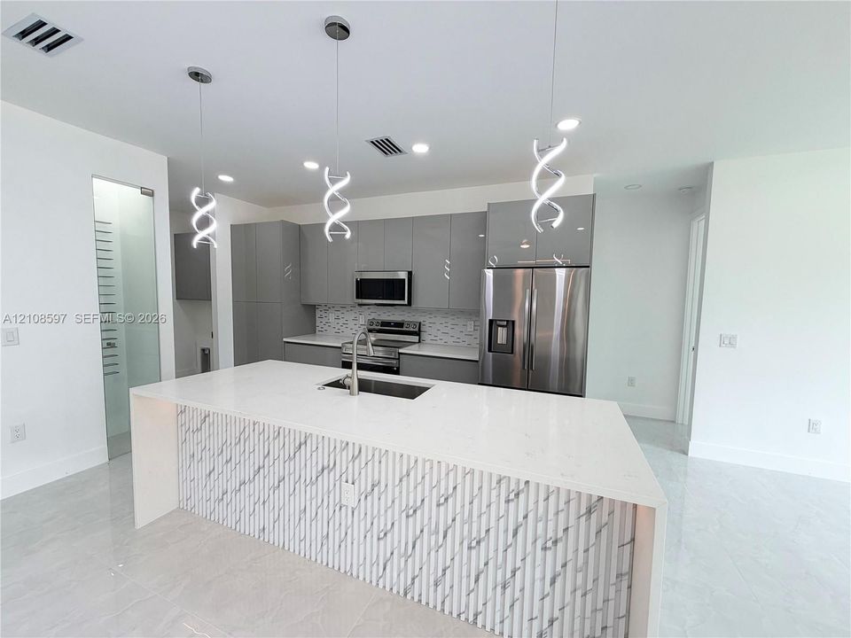 Kitchen featuring high-gloss flat-panel cabinetry, a waterfall-edge island with stone-surface countertop, tile backsplash, stainless steel appliances, and sculptural pendant lighting