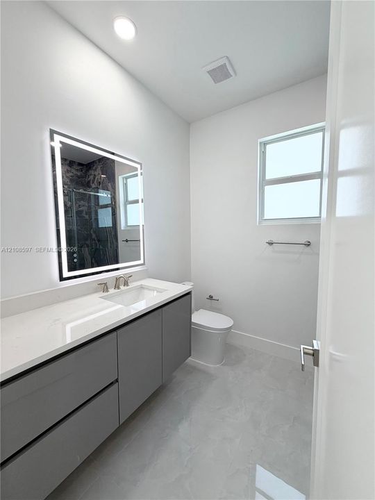 Modern vanity featuring a stone countertop, integrated sink, and illuminated rectangular mirror, complemented by grey cabinetry, light-toned flooring, and a window for natural light