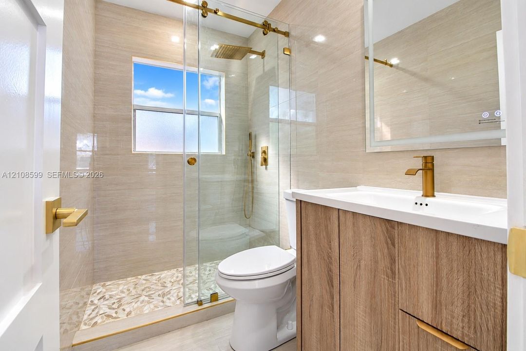 Bathroom featuring a wood-finish vanity with a gold-tone faucet, a glass-enclosed shower with gold-tone hardware, textured tile wall surrounds, and a built-in illuminated mirror