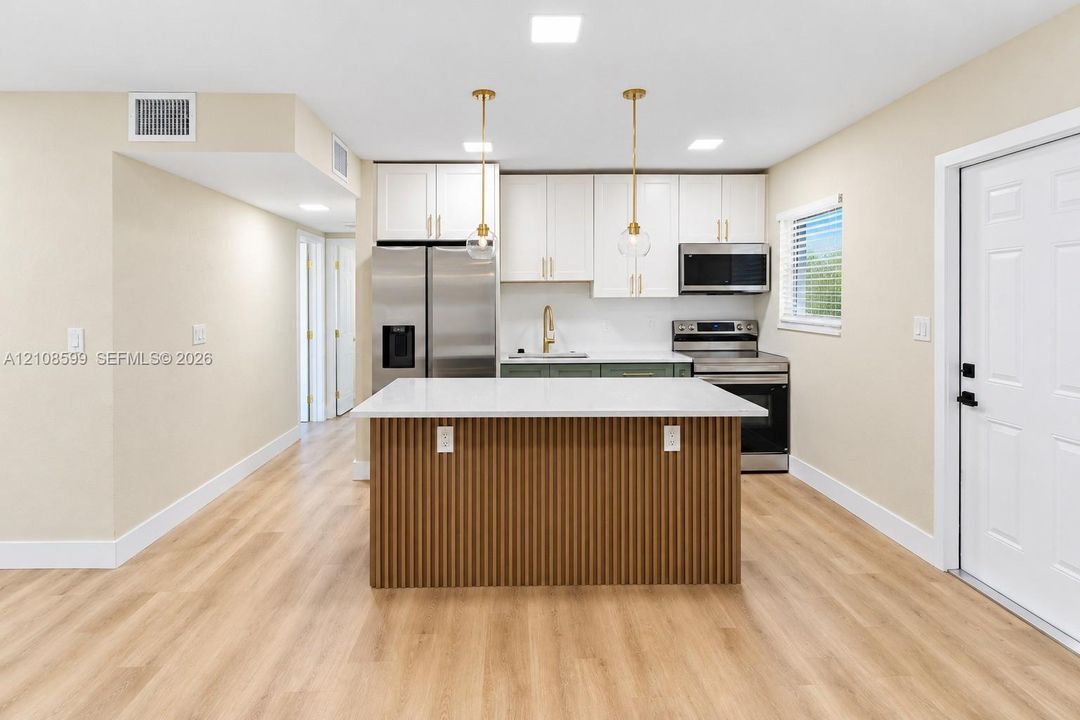Kitchen featuring white shaker-style cabinetry, stainless steel appliances, a center island with a ribbed wood-finish base and light-toned countertop, and dual brass pendant lights