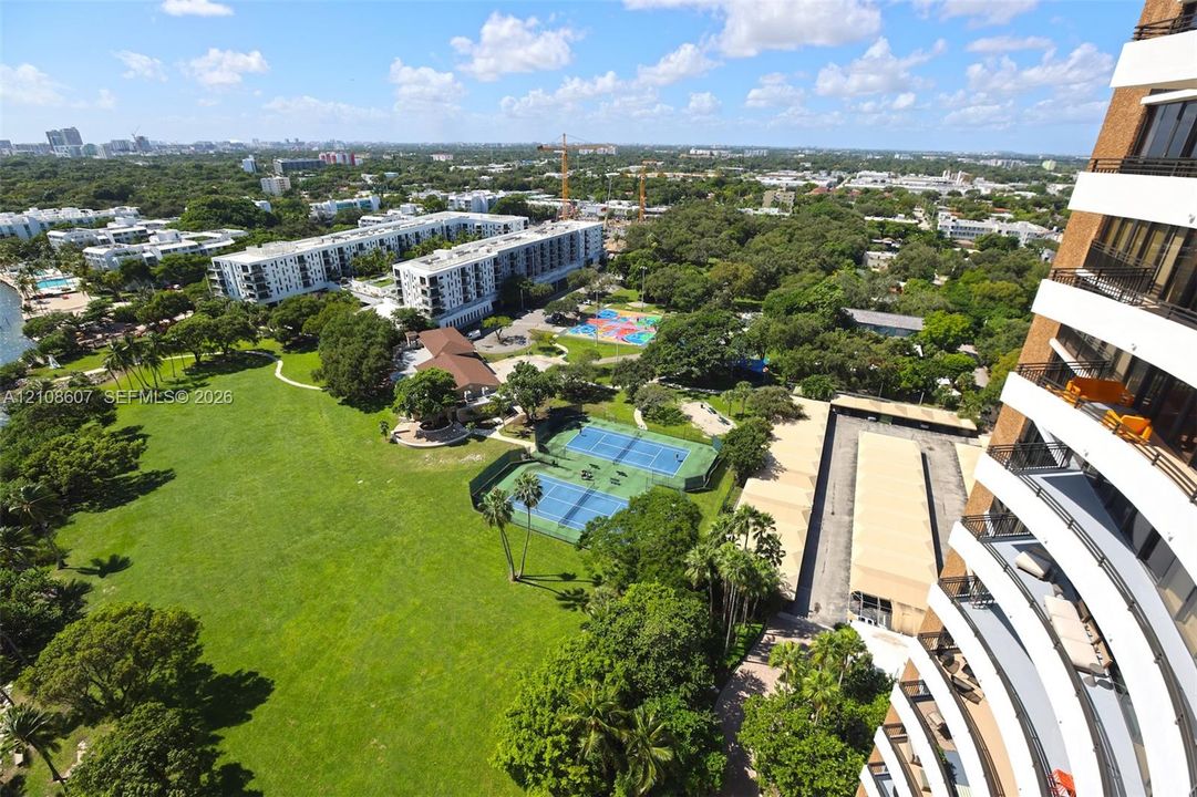 High-rise residential exterior with stacked cantilevered balconies, dual tennis courts, expansive green space, and covered parking structures
