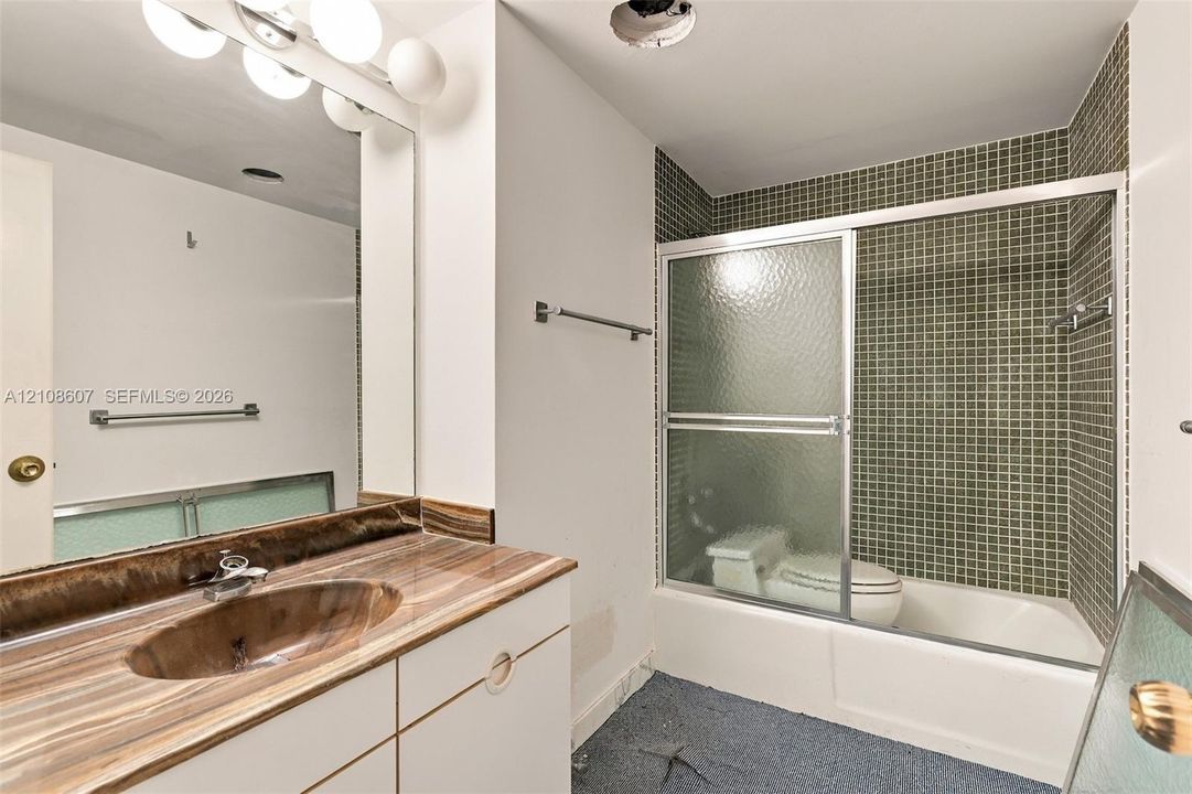 Vanity featuring a natural stone-patterned countertop with a recessed basin, integrated wall-width mirror, shower and tub combination with glass sliding doors, and green mosaic tile surround