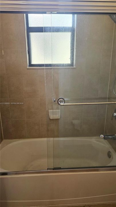 Bathtub and shower combination featuring neutral-toned wall tile, a sliding glass door enclosure, built-in soap niche, and a window with privacy glass