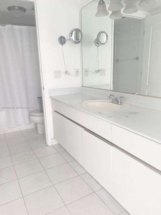 Bathroom featuring a white vanity with a solid surface countertop, an integrated sink, dual wall-mounted magnifying mirrors, a large wall-to-wall mirror, and ceramic tile flooring