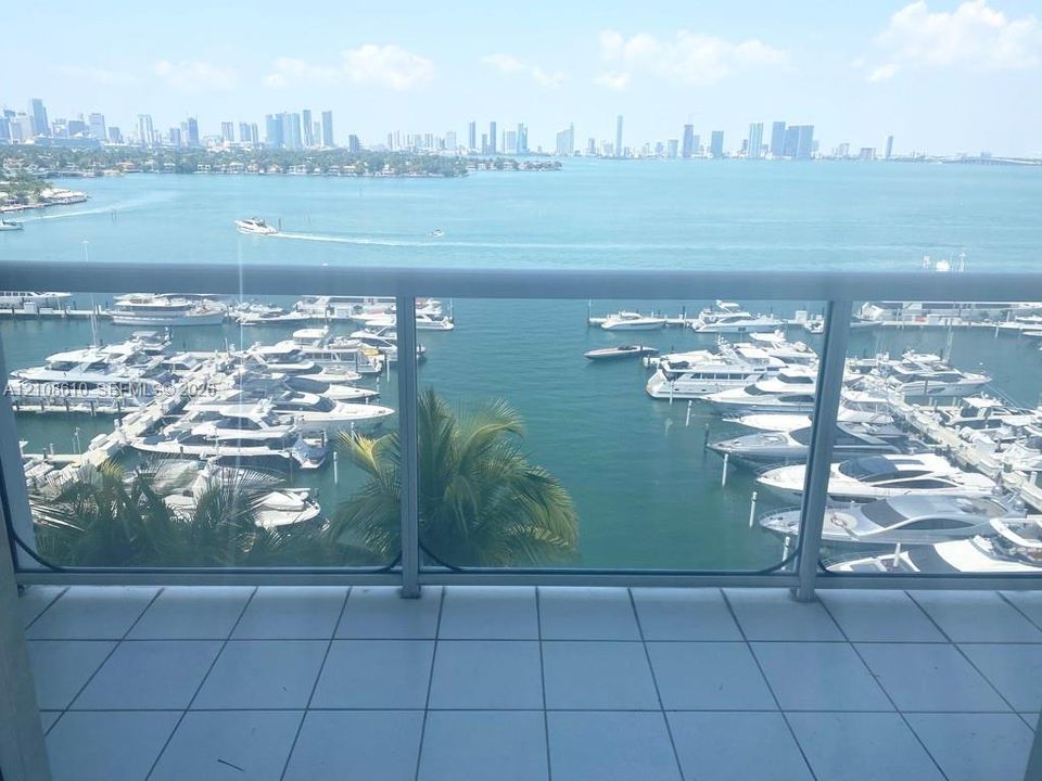 Private balcony featuring white tile flooring and a glass-paneled railing, overlooking a marina and city skyline