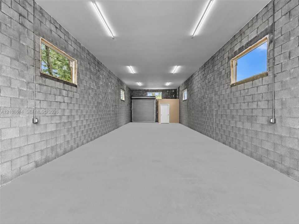 Expansive workspace featuring concrete block walls, a smooth concrete floor, multiple windows, overhead lighting, and a metal roll-up door