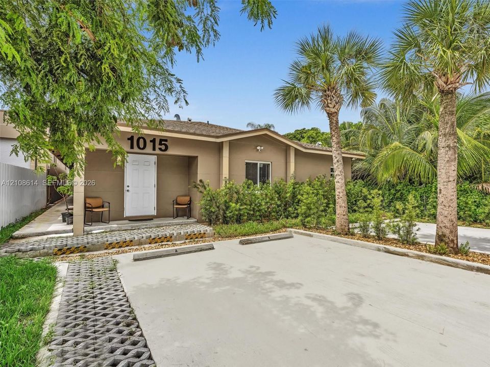Single-story exterior featuring a covered entryway, concrete paved parking area, perimeter landscaping, and mature palm trees