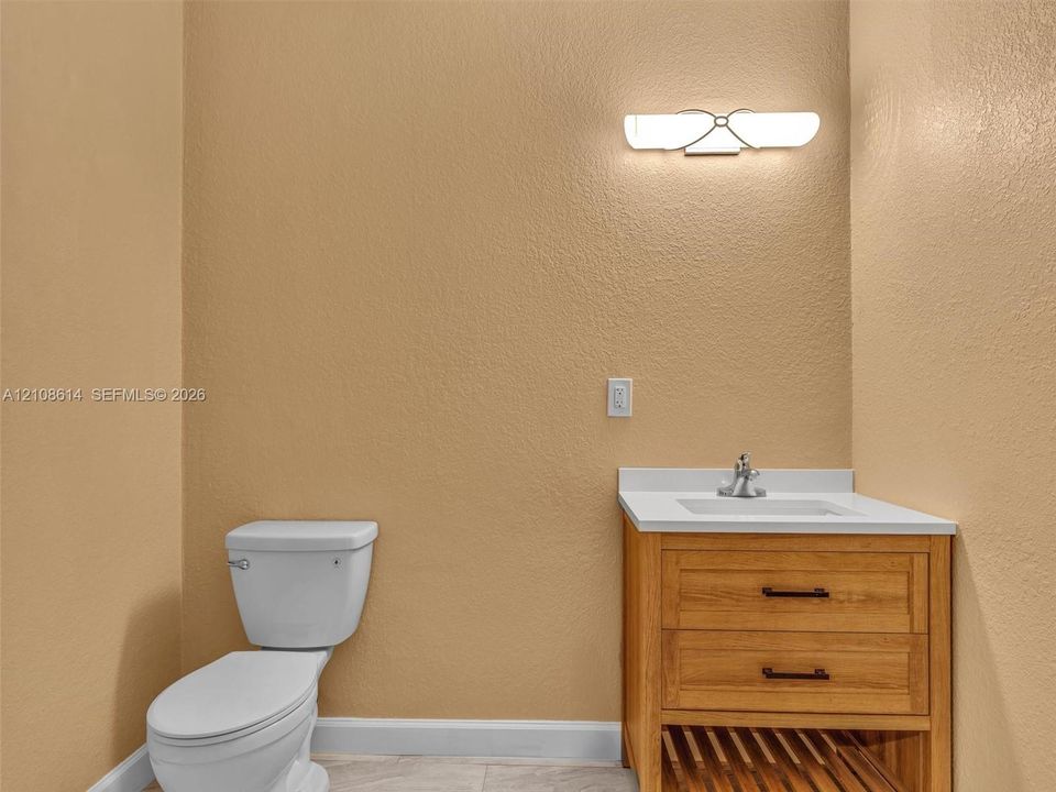 Wood-finish vanity with white countertop, integrated sink, chrome faucet, and dual-drawer storage featuring matte black hardware, accompanied by a wall-mounted light fixture and high-profile toilet