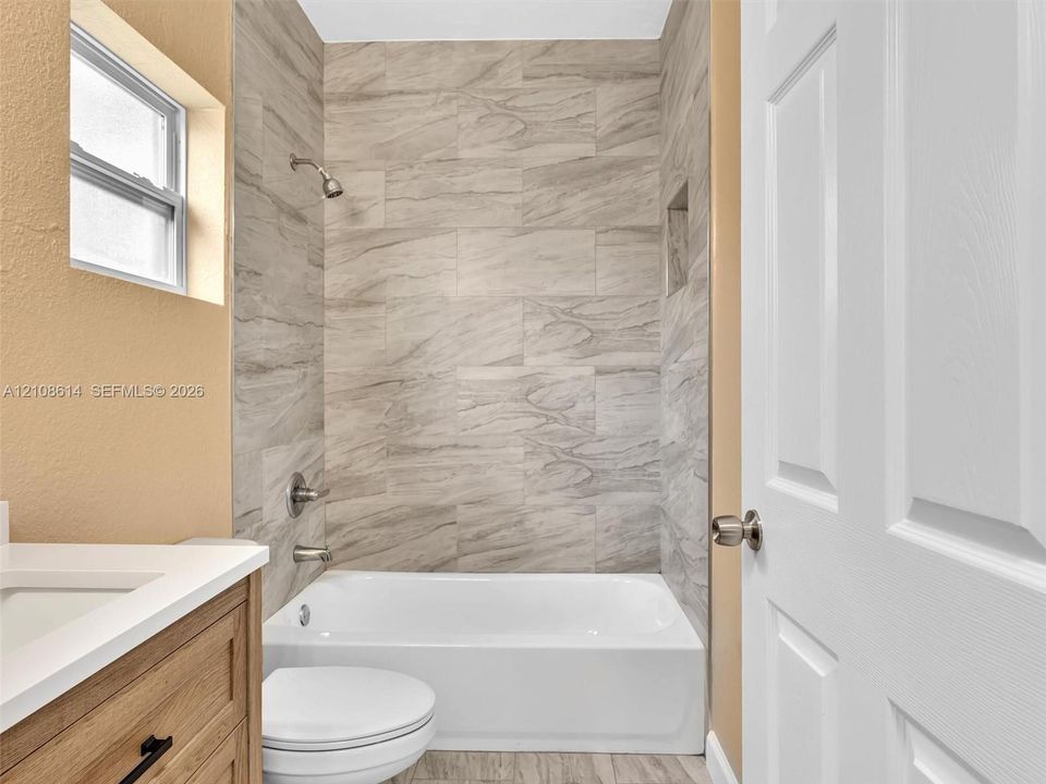 Bathroom featuring a tub-shower combination with floor-to-ceiling stone-look tile surround, built-in wall niche, wood-finish vanity with a rectangular sink, and polished metal fixtures