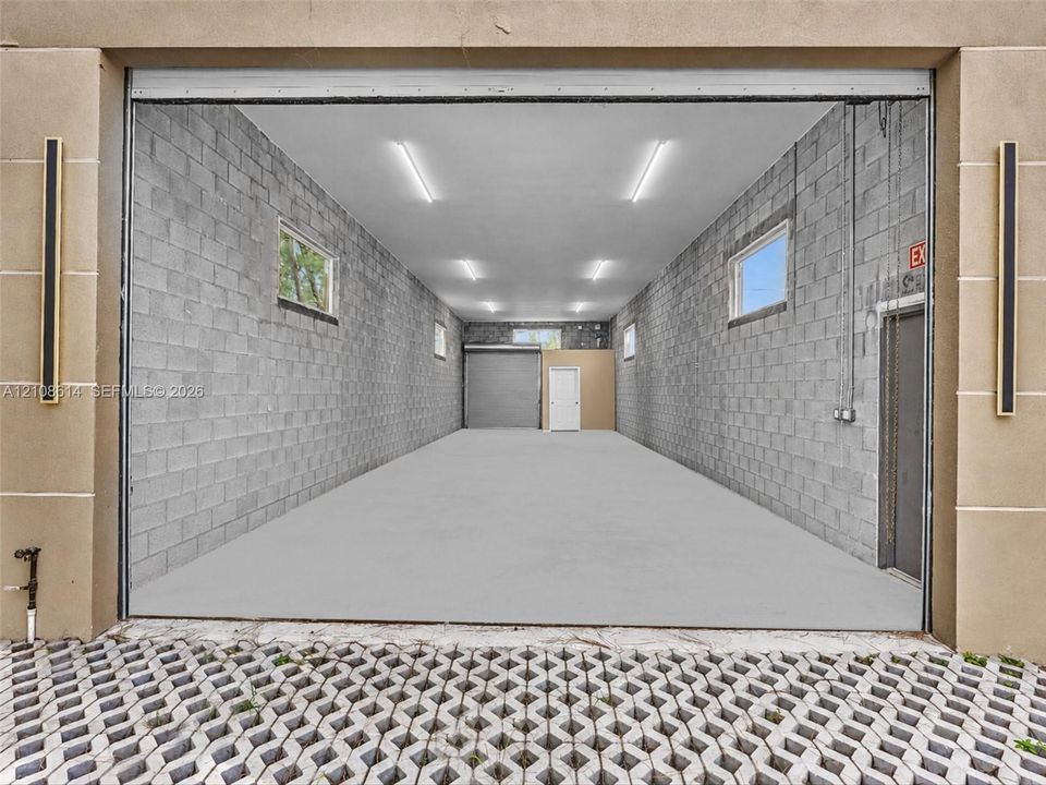 Expansive garage featuring concrete block walls, overhead lighting, multiple windows, a secondary roll-up door, and an exterior permeable paver apron