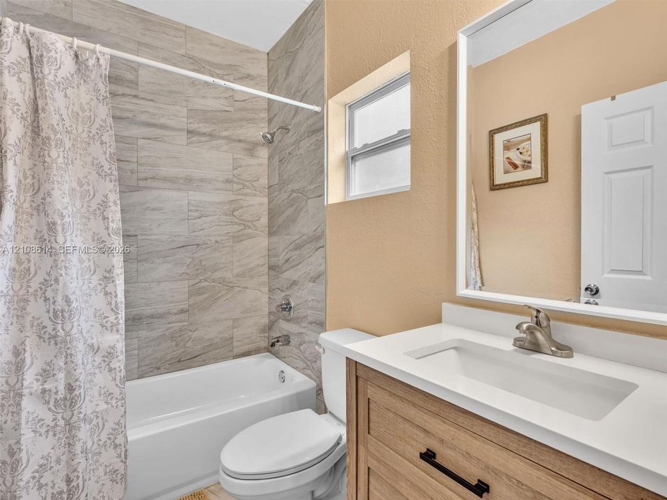 Bathroom featuring a vanity with a light wood finish, white countertop, integrated rectangular sink, and matte black hardware, accompanied by a tub enclosure with neutral-toned horizontal wall tile