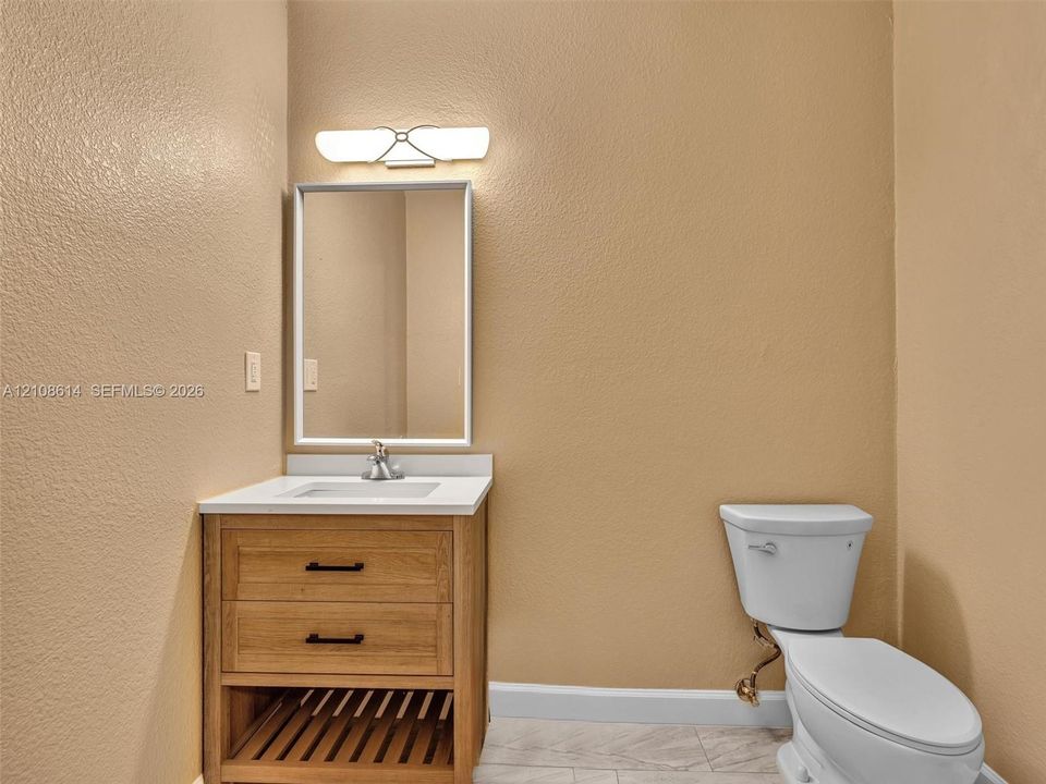Wood-finish vanity with a rectangular sink, white countertop, matte black hardware, and an integrated slatted lower shelf, paired with a modern framed mirror, two-light fixture, and neutral tile flooring