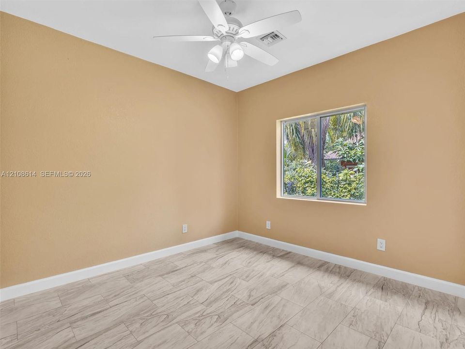 Neutral-toned interior space featuring wood-finish flooring, a white ceiling fan with integrated lighting, and a window with exterior greenery
