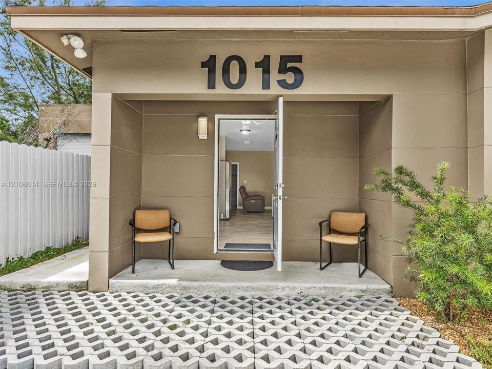 Covered entryway featuring a concrete stoop, geometric permeable paver driveway, wall-mounted exterior lighting, and stucco-style facade
