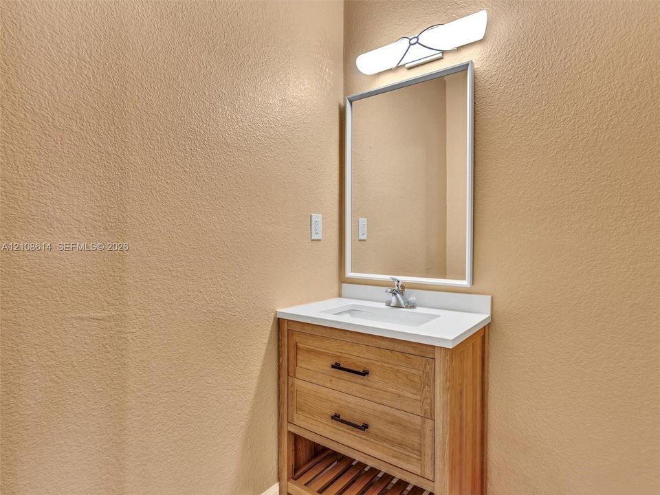 Wood-finish vanity featuring dual drawers, an open-slatted lower shelf, a white rectangular countertop with an integrated basin, and a framed mirror paired with a linear wall-mounted light fixture