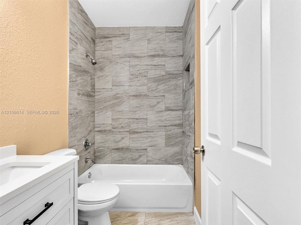 Bathroom featuring a white vanity cabinet with a stone-surface countertop, tub-shower combination with full-height neutral-toned tile surround, built-in wall niche, and six-panel interior door