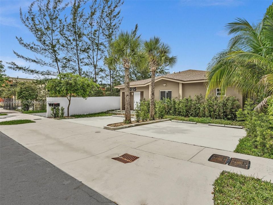 Exterior facade featuring a neutral-toned finish, shingled roof, concrete driveway, white privacy fencing, and mature palm trees
