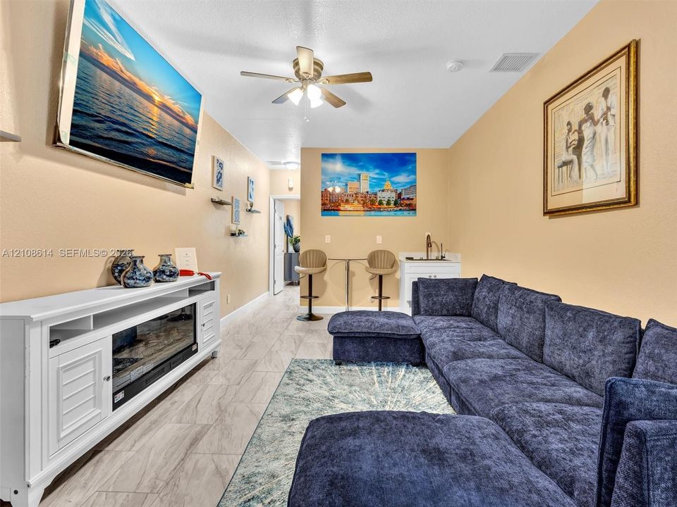 Built-in white media cabinet with integrated electric fireplace, wood-finish tile flooring, ceiling fan with light fixture, and kitchenette wet bar with sink