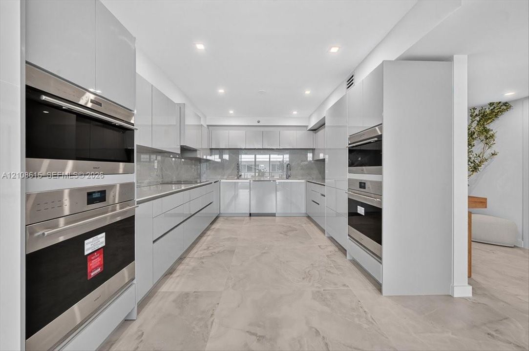 Contemporary kitchen featuring high-gloss cabinetry, stainless steel double wall ovens, integrated appliances, stone-finish backsplash, and large-format neutral tile flooring