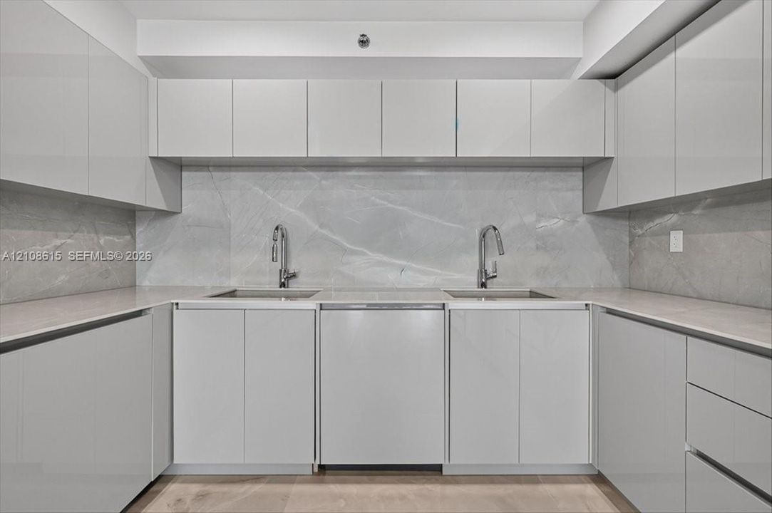 Glossy cabinetry, stone slab backsplash, dual undermount sinks, gooseneck faucets, and light-toned countertop surfaces