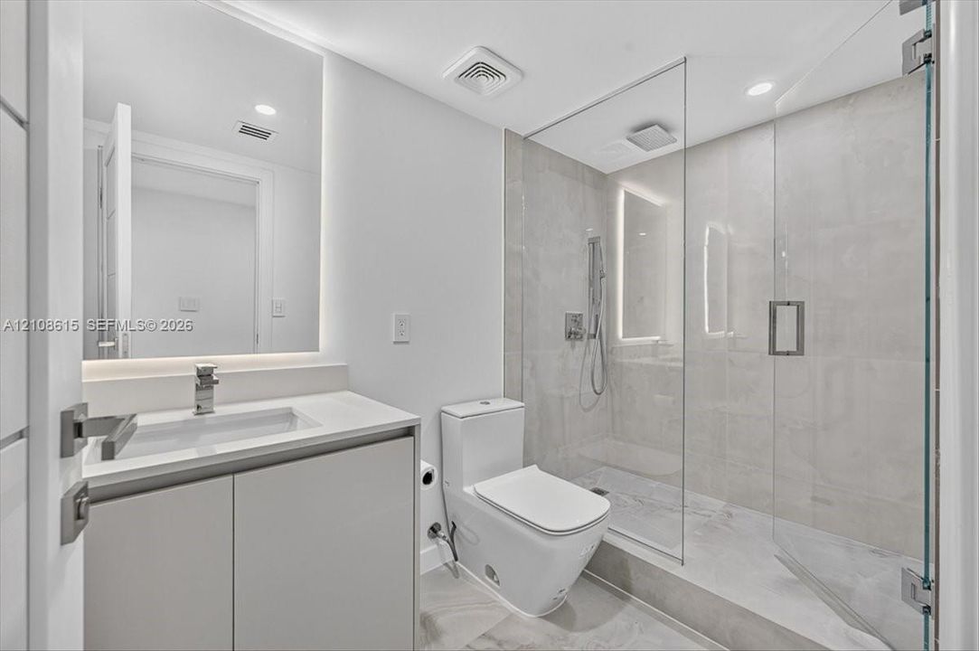Modern bathroom featuring a floating vanity with a light-toned countertop, a seamless walk-in shower with a glass enclosure, large-format tile flooring, and backlit wall mirror