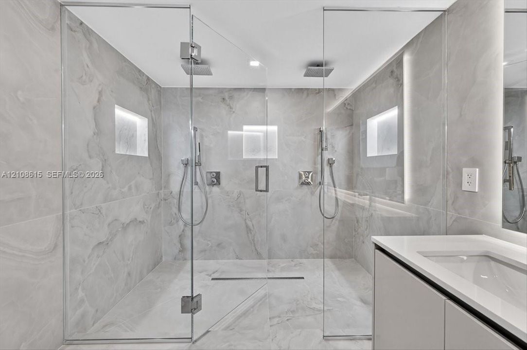 Walk-in shower featuring floor-to-ceiling stone-finish tile, dual ceiling-mounted rainfall showerheads, handheld sprayers, recessed wall niches, and frameless glass enclosure