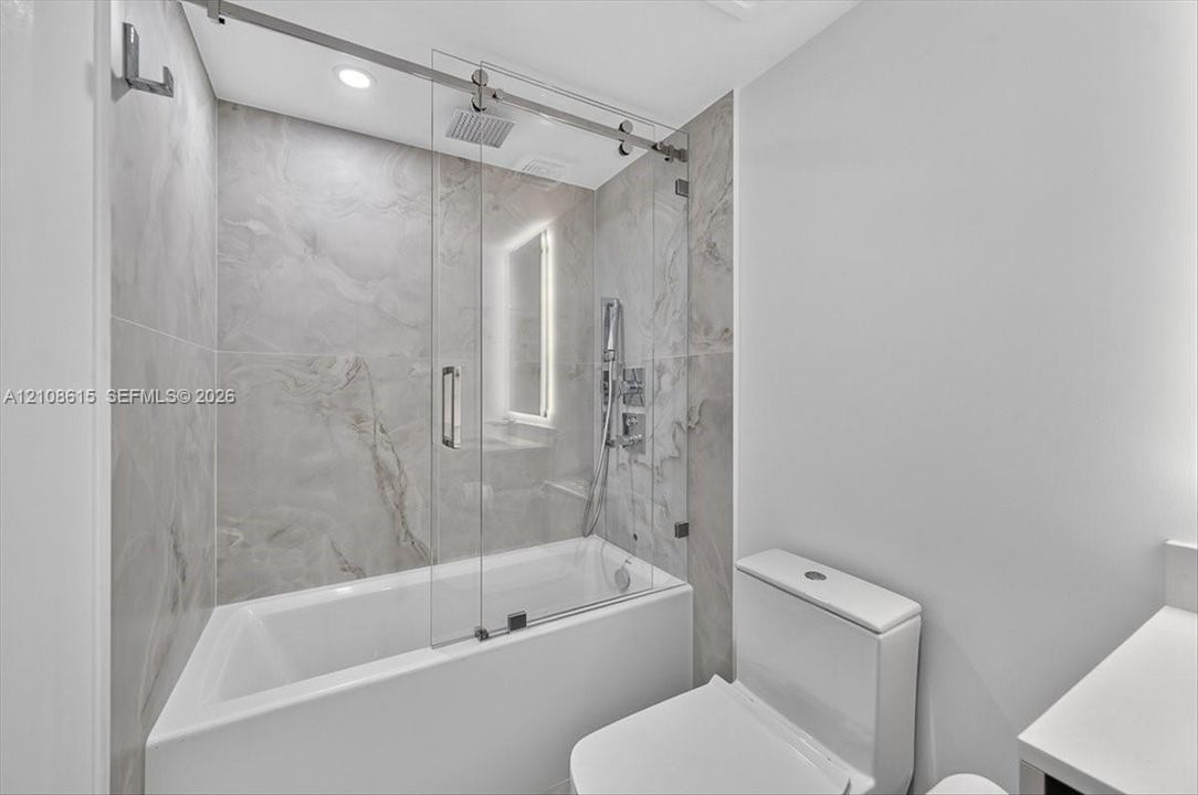 Shower with glass sliding enclosure, floor-to-ceiling stone-finish tile surround, ceiling-mounted rainfall showerhead, and integrated handheld fixture