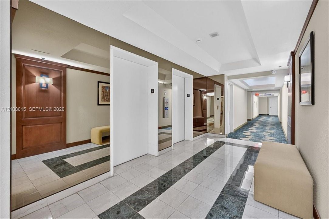 Elevator lobby featuring floor-to-ceiling wall mirrors, polished stone tile flooring with decorative inlay, and wood-paneled accents