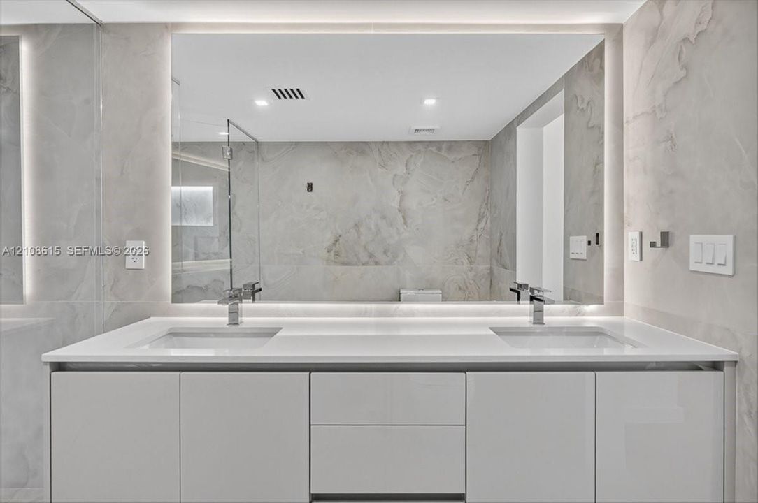 Modern dual-sink vanity featuring a solid-surface countertop, high-gloss cabinetry, integrated LED-backlit mirror, and polished metal fixtures against full-height stone-finish wall tile
