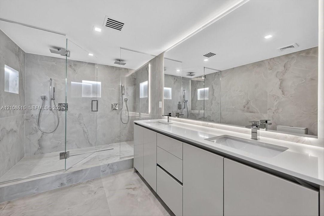 Bathroom featuring a walk-in shower with glass enclosure, large-format tile surround, dual vanity with undermount sinks, full-width mirror, and integrated lighting