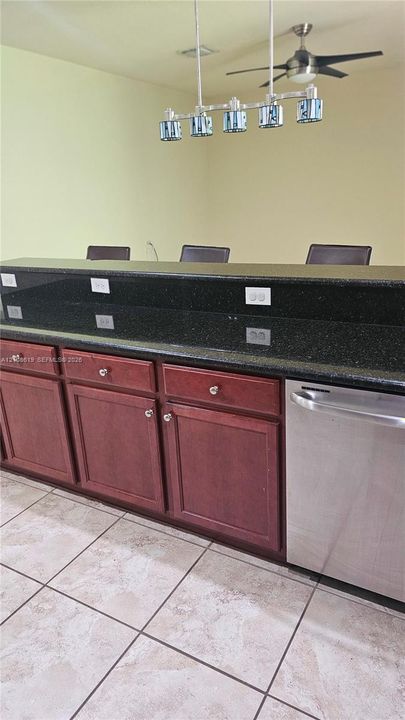 Kitchen island featuring a dark stone countertop, stained wood cabinetry, stainless steel dishwasher, five-light pendant fixture, and ceramic tile flooring