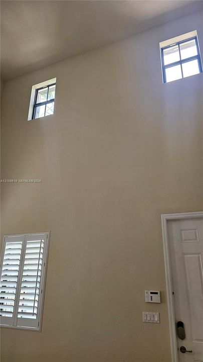 High-ceiling interior featuring dual clerestory windows, a lower window with white louvered plantation shutters, and a six-panel door with architectural molding