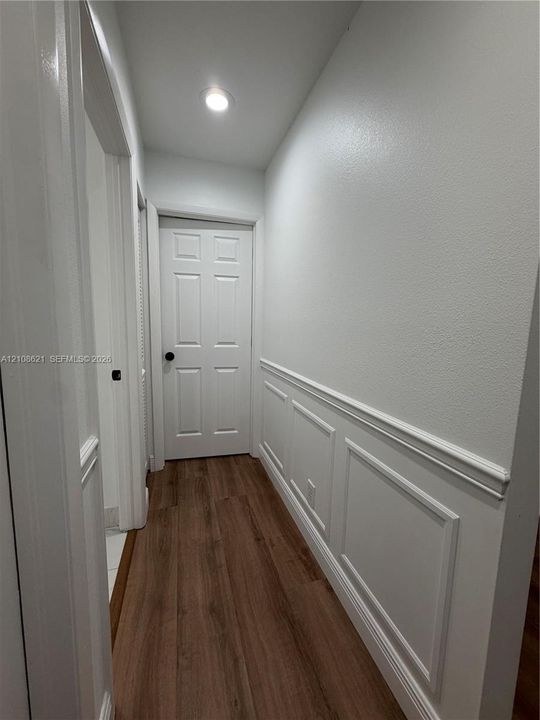 Hallway featuring wood-finish flooring, white paneled wainscoting, recessed ceiling lighting, and a six-panel door with a matte black lever handle