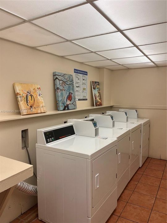 Laundry facility featuring multiple white front-loading appliances, a built-in wall shelf, a drop ceiling with integrated lighting, and tile flooring