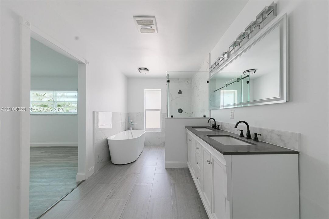 Dual-sink vanity with a dark countertop and matte black fixtures, freestanding soaking tub, glass-enclosed shower with tiled surround, and large-format tile flooring