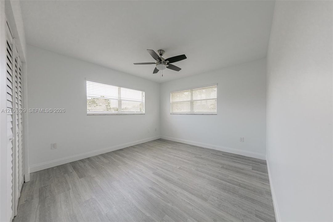 Neutral-toned room featuring wood-finish flooring, dual windows with horizontal blinds, a ceiling fan, and louvered closet doors