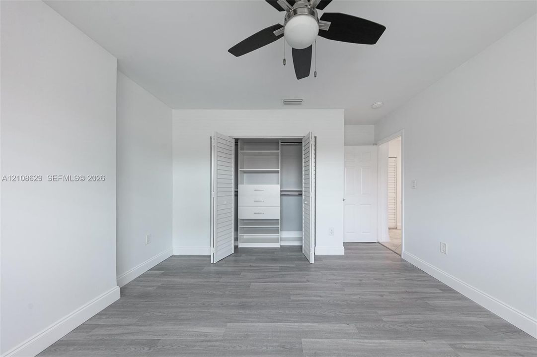 Neutral-toned interior featuring wood-finish flooring, a ceiling fan with integrated lighting, and a closet with white louvered doors and built-in shelving and drawer units